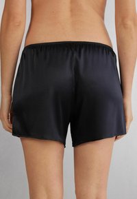 Zwarte satijnen shorts met een gladde textuur, elastische tailleband en een losse pasvorm, tot midden boven de dijen gesneden. Geen zichtbare patronen of accenten.