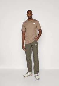 Ruda medvilninė marškinėliai su logotipu ant krūtinės, derinami su alyvuogių žalių krovinių kelnių su kišenėmis ir diržo kilpomis, dėvimi su šviesiais sportiniais batais.