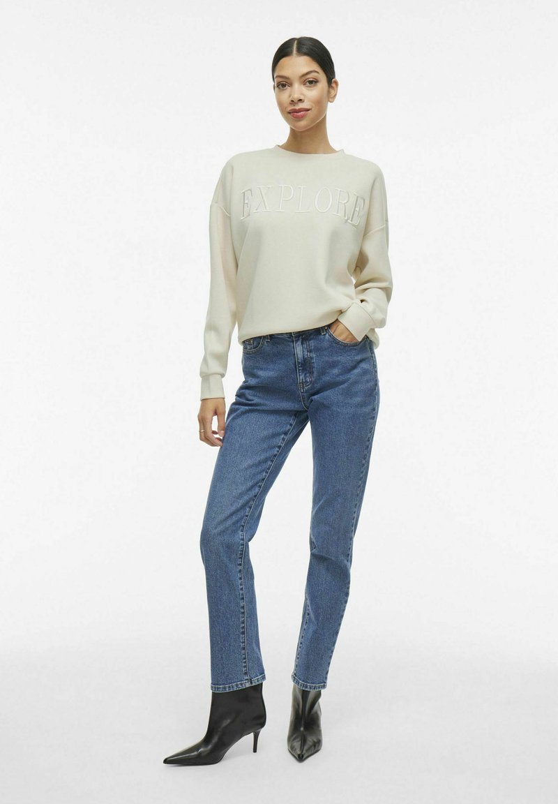 Hellbeige Sweatshirt mit geprägtem Text "EXPLORE", kombiniert mit blauen, hochgeschnittenen Jeans und schwarzen, spitzen Knöchelstiefeln.