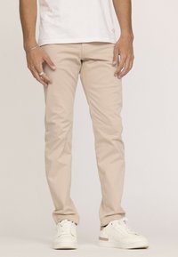 Pantalon en coton beige avec une coupe slim, texture lisse et poches avant, associé à des baskets blanches avec une subtile touche dorée.