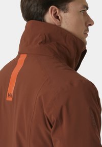 Hnědá nepromokavá bunda s vysokým límcem, oranžovým vertikálním akcentem a značkováním RECCO na rukávu. Hladká textura a aerodynamický design.
