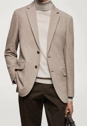 Mann trägt einen beigen Blazer über einem hellbeigen Rollkragenpullover und dunkelbraune Hose, hält einen braunen Lederriemen in der rechten Hand.