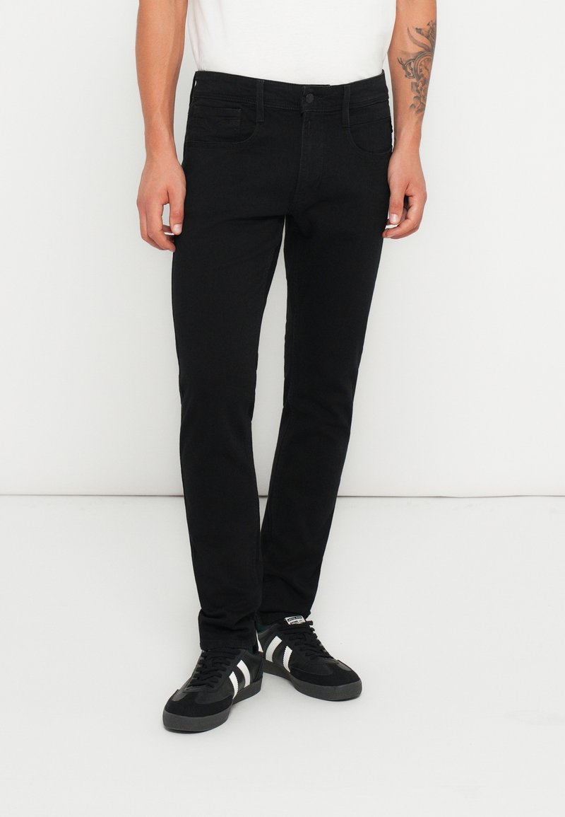 Homme portant un jean noir slim et des baskets noires et blanches, debout contre un mur blanc.