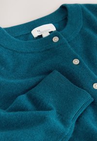 Petrolejno modra volnena jopica z okroglim izrezom, zapiranjem na gumbe s svetlimi gumbi in rebrastimi manšetami, ki izkazuje mehko teksturo.