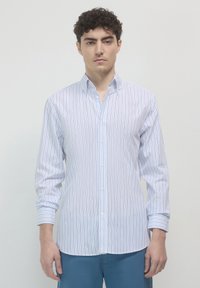 Junger Mann mit kurzen lockigen Haaren, der ein weißes Hemd mit schmalen blauen vertikalen Streifen und blaue Hosen trägt, vor einem einfarbigen Hintergrund stehend.