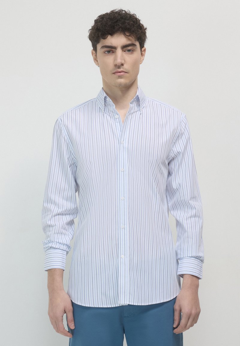 Junger Mann mit kurzen lockigen Haaren, der ein weißes Hemd mit schmalen blauen vertikalen Streifen und blaue Hosen trägt, vor einem einfarbigen Hintergrund stehend.