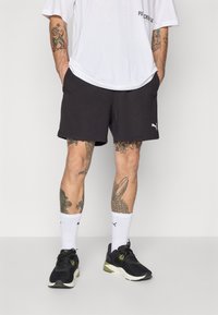 Shorts de sport Puma en gris foncé, associés à des baskets noires et olive, des chaussettes blanches à mi-mollet et un t-shirt blanc ample. Tatouages notables visibles.