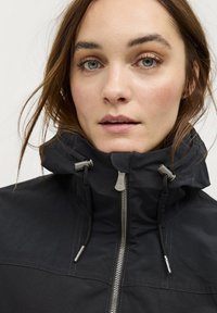 Veste noire imperméable avec un col montant, une fermeture éclair à l'avant et des cordons de serrage réglables. Présente du matériel métallique et une texture lisse.