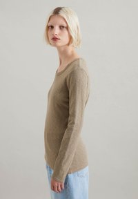 Long-sleeve shirt in a soft beige material with a rounded neckline and fitted silhouette, paired with light blue denim jeans.