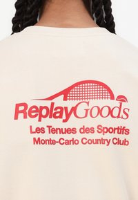 Light beige sweatshirt featuring bold red text that reads “Replay Goods” with a graphic of a tennis racket and net, plus additional smaller text below.