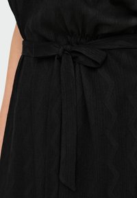 Črna obleka z teksturirano tkanino, ki ima pas z navezanim vozlom. Material ima valovito vzorčenje, ki poudarja globino in detajle.