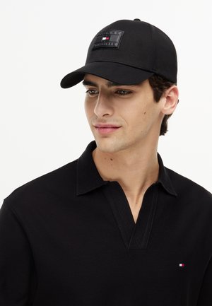 Jonge man die een zwarte pet met Tommy Hilfiger-logo draagt en een zwart overhemd met kraag met een klein Tommy Hilfiger-embleem op de borst.