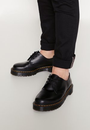 Personne portant des chaussures en cuir noir à lacets avec des semelles épaisses et des coutures jaunes, associées à un pantalon noir retroussé.