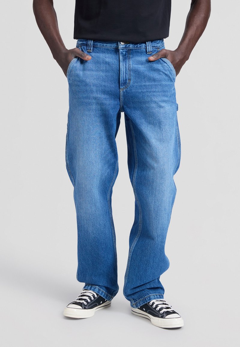 Homme portant un jean bleu larges, les mains dans les poches, et des baskets noires et blanches sur un fond gris uni.
