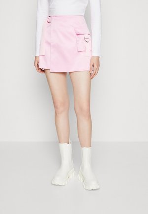 Femme portant une mini-jupe portefeuille rose avec détail de poche latérale, un haut blanc à manches longues et des bottines blanches épaisses, debout contre un mur blanc.