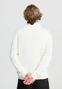 Maglione bianco lavorato a maglia con collo alto a coste e polsini. Texture morbida con un design aderente, visto da dietro su uno sfondo grigio.