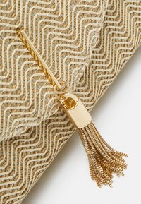 Gland métallique dorée attachée à un tissu beige tissé texturé avec un motif géométrique, photographiée sur fond blanc.