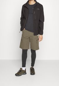 Chaqueta negra con cremallera, camiseta gris, pantalones cortos cargo verde oliva, mallas negras y zapatos de senderismo marrones. Cremalleras y texturas destacadas.