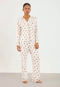 Vrouw die blootsvoets staat, gekleed in een witte, lange mouwen, knoopsluiting pyjama en broek met een bruin teddybeer patroon.