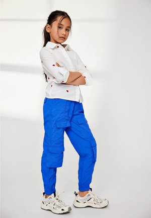 Camicia bianca con motivi a cuore rossi, pantaloni cargo blu con polsini elasticizzati, abbinati a sneaker bianche con accenti neri.