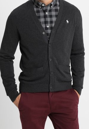 Man wearing dark gray buttoned cardigan over black and gray checkered shirt with burgundy pants, hands in pockets.