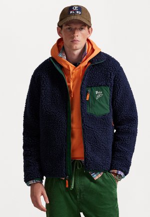 Jeune homme portant une casquette marron, un sweat à capuche orange, une veste en sherpa marine avec une poche verte, une chemise à carreaux, un pantalon en velours côtelé vert et une montre.