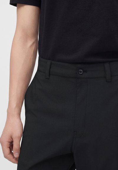 Homme portant un pantalon noir et une chemise noire, avec un focus sur la taille et le bras gauche sur un fond uni.