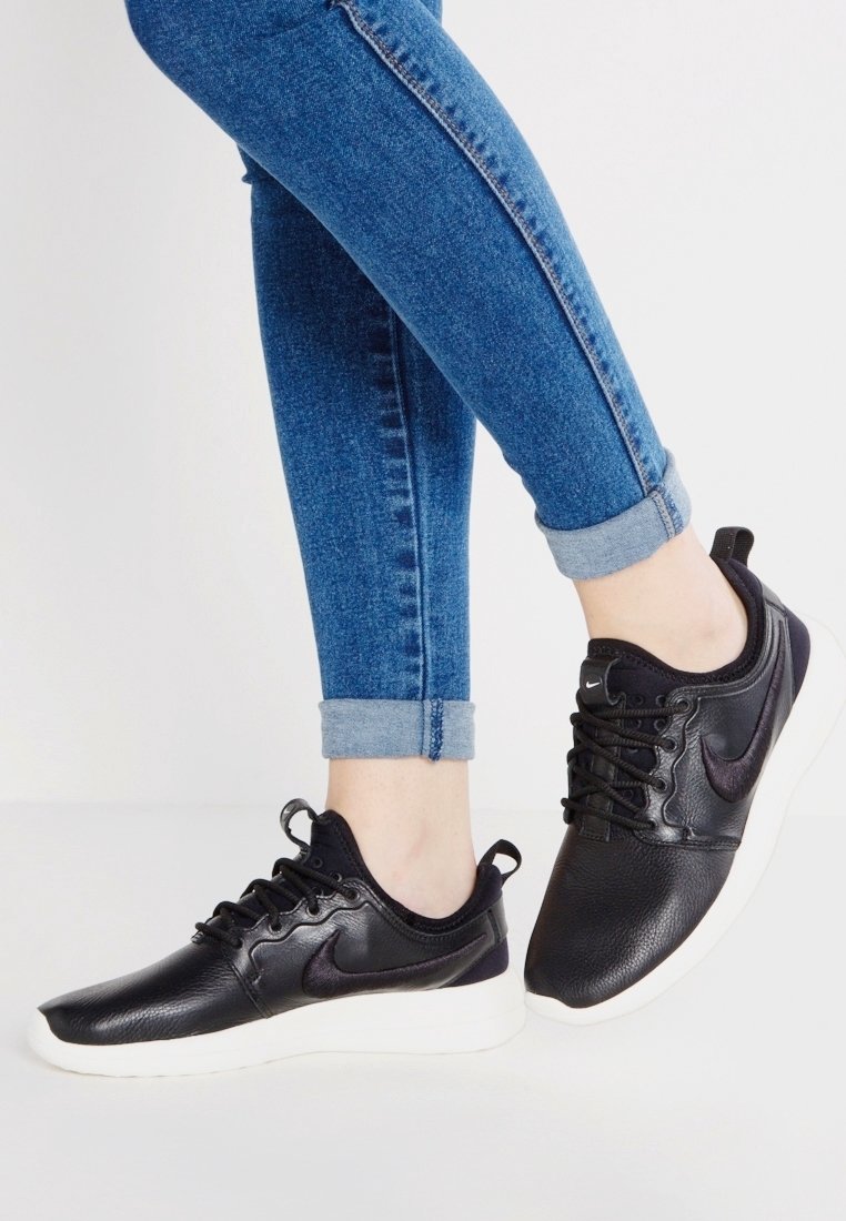 Personne portant un jean bleu retroussé et des baskets Nike noires avec semelles blanches, debout sur un sol blanc devant un fond uni.