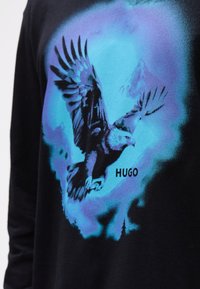 Cămașă neagră cu mâneci lungi, prezentând un grafic cu un vultur în tonuri de albastru și teal, cu cuvântul "HUGO" imprimerat dedesubt.