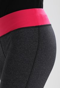 Detail osoby oblečené v tmavě šedých legínách se širokým jasně růžovým páskem v pase a černém topu.