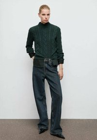 Suéter de punto trenzado verde oscuro con cuello alto, combinado con jeans de mezclilla oscuros y sueltos y tacones negros puntiagudos. Diseño simple y texturizado.