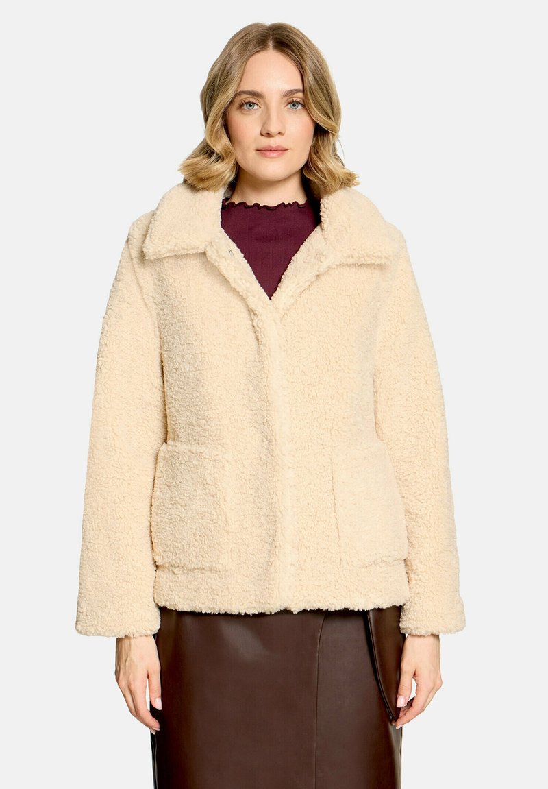 Femme portant une veste polaire beige texturée par-dessus un haut bordeaux et une jupe en cuir marron, debout devant un fond clair uni.