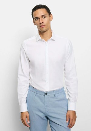 Uomo che indossa una camicia bianca a maniche lunghe con bottoni infilata in pantaloni blu chiaro, in piedi davanti a uno sfondo bianco uniforme.