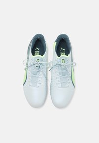 Zapatos de fútbol sintéticos blancos con acentos en azul claro, parte superior texturizada, cordones planos y tacón azul oscuro, con una franja en verde neón.