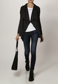 Svart, draperad cardigan i texturerat tyg, lagd över en vit topp. Bärs med mörka denimjeans och svarta ankelstövlar, håller en svart väska.