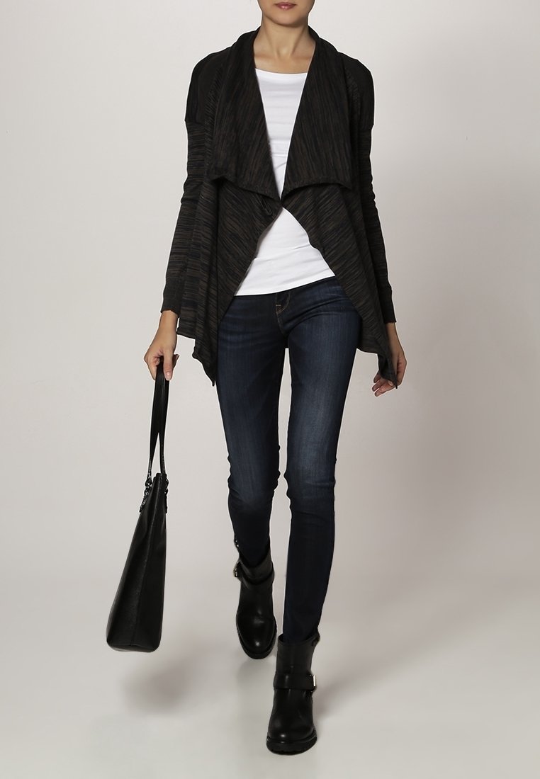 Svart, draperad cardigan i texturerat tyg, lagd över en vit topp. Bärs med mörka denimjeans och svarta ankelstövlar, håller en svart väska.