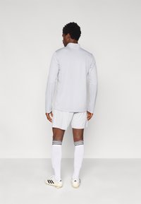 Αθλητική μπλούζα ανοιχτού γκρι με μακριά μανίκια, ψηλό γιακά, συνδυασμένη με αντίστοιχα σορτς. Άσπρες κάλτσες με μαύρες ρίγες και ποδοσφαιρικά παπούτσια.