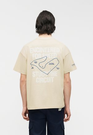 Homme aux cheveux foncés portant un t-shirt beige montrant une carte de circuit de course avec le texte "Conçu pour la vitesse Étudiez le circuit" au dos.