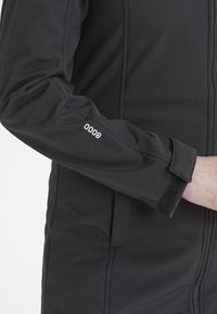 Musta pehmeäkuorijakku, jossa on vetoketjukiinnitys, teksturoitu kangas, säädettävät hihansuut tarranauhalla ja "8000 WPRO" -brändäys hihassa.