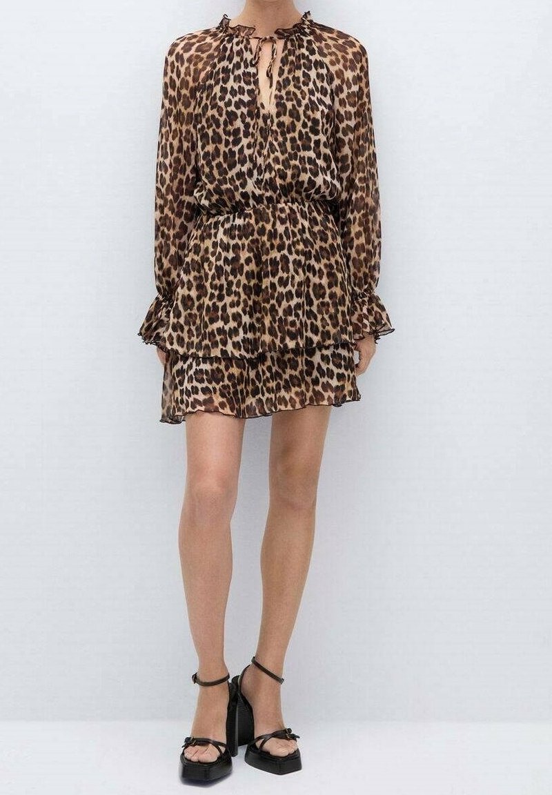 Femme portant une mini-robe à manches longues imprimée léopard associée à des sandales noires à talons hauts avec brides à la cheville sur un fond uni.