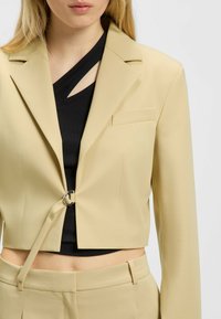 Korte beige blazer met een enkele knoopsluiting met een zilveren ring, gecombineerd met een zwarte ribbeltop met een uitsnijding.