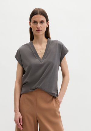 Vrouw met steil bruin haar, die een grijs T-shirt met V-hals en korte mouwen draagt en een camelgekleurde broek met hoge taille, met één hand in de zak.