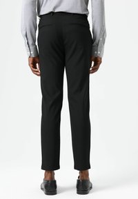 Zwarte broek met een getailleerde pasvorm, voorzien van achterzakken en een gladde textuur. Gecombineerd met zwarte schoenen en een lichtgrijs overhemd.