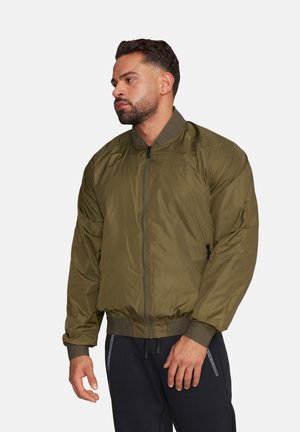 Mężczyzna o krótkich ciemnych włosach i brodzie ubrany w oliwkową kurtkę typu bomber oraz czarne spodnie, stoi na jasnym tle, patrząc w lewo.