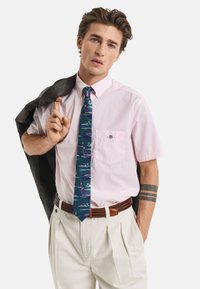 Korte mouwen roze overhemd met knopen, blauwe gestreepte stropdas, lichtbeige geplooide broek, bruine gevlochten riem, leren jack over één schouder gedragen.