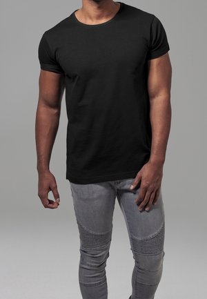 Homme portant un t-shirt noir près du corps à manches courtes et un jean gris clair côtelé, debout devant un fond gris uni.