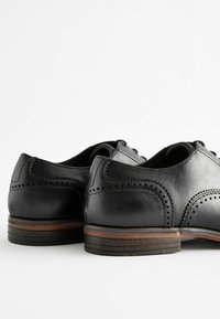 Zapatos de cuero negro con detalles de brogue, punta redonda y cordones de cuero. Suela de madera con una ligera elevación y acentos de costura en contraste.