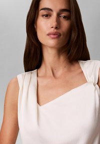 Femme aux longs cheveux bruns portant un haut blanc texturé sans manches avec un décolleté carré, regardant la caméra avec confiance sur un fond gris.
