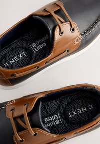 Tige en cuir marron avec accents bleu marine, semelle en caoutchouc blanc et surpiqûres décoratives. Équipée de semelles intérieures Ortholite® Ultra pour le confort.