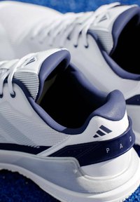 Bílé a námořnické sportovní boty s texturovaným síťovaným svrškem, polstrovaným límcem a výrazným brandingem Adidas na boku a patě.